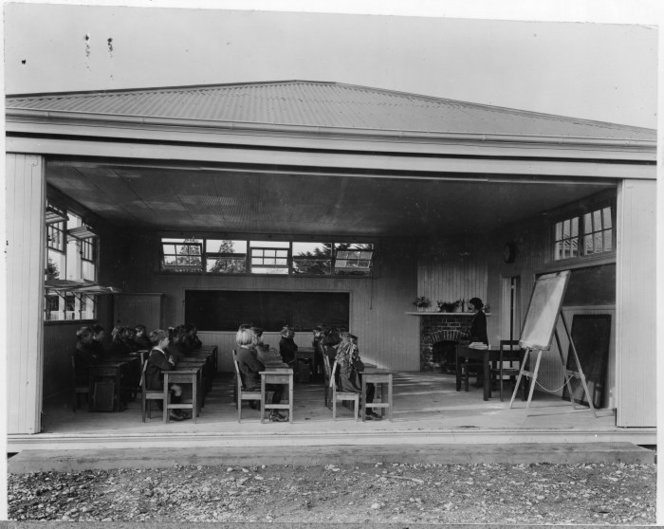 Photograph of an open air classroom, Fendalton School, Christchurch, taken circa 1924 by an unidentified photographer. Primary school children sit in rows at their desks, facing a teacher and a blackboard.